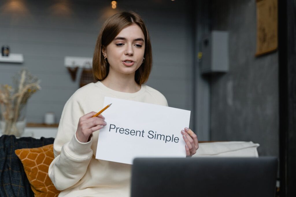pexels photo 6671683 Young woman teaches English grammar online, holding a 'Present Simple' note and pencil.