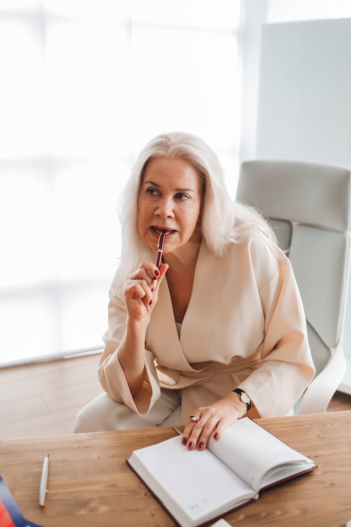 Woman in office thinking with pen, open book on desk, stylish setting.